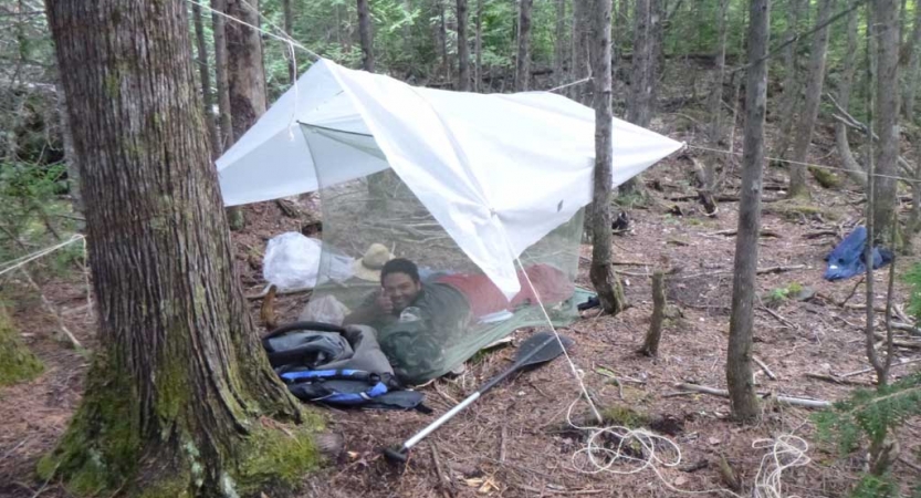 a person smiles from under a tarp shelter amongst trees on an outward bound course